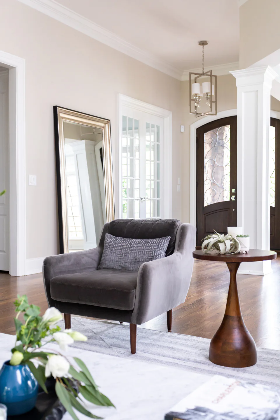 Gray velvet armchair with a patterned pillow beside a turned wood side table, white tulips in the foreground, in the living room at Durham Transitional, Durham, North Carolina, interior design by Alexandra Peck Design