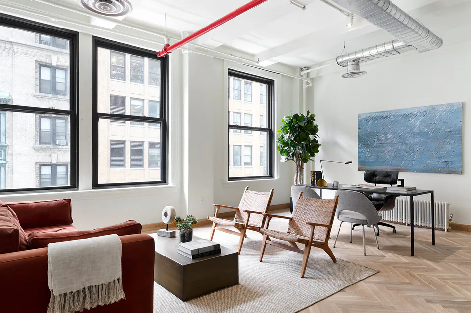 Executive office with a rust velvet sofa, woven lounge chairs, a desk and a large blue painting beneath steel-framed windows and exposed ductwork at Flatiron Workplace, 3 East 28th Street, New York City, interior design by Alexandra Peck Design