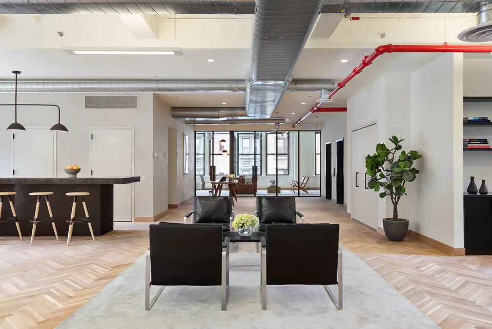 Open lounge with black leather lounge chairs on a pale rug, the pantry bar and exposed ductwork with red sprinkler pipes at Flatiron Workplace, 3 East 28th Street, New York City, interior design by Alexandra Peck Design