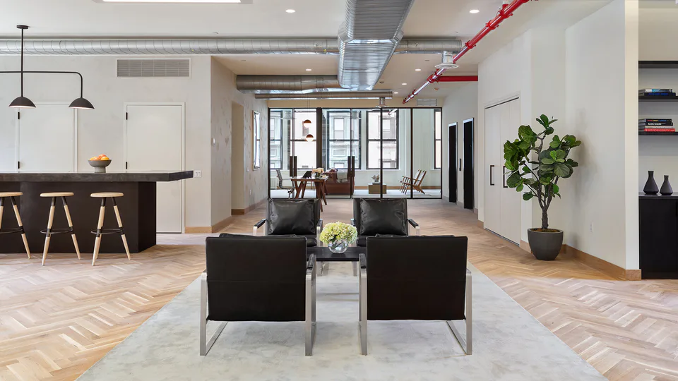 Open lounge with black leather lounge chairs on a pale rug, the pantry bar and exposed ductwork with red sprinkler pipes at Flatiron Workplace, 3 East 28th Street, New York City, interior design by Alexandra Peck Design