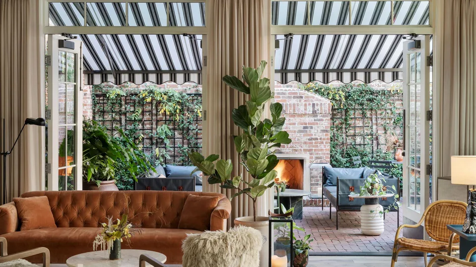 Hotel lobby with a tufted terracotta sofa and shearling chairs, its doors open to the garden courtyard under striped awnings, at The Pinch Hotel, Charleston, South Carolina, interior design by Alexandra Peck Design
