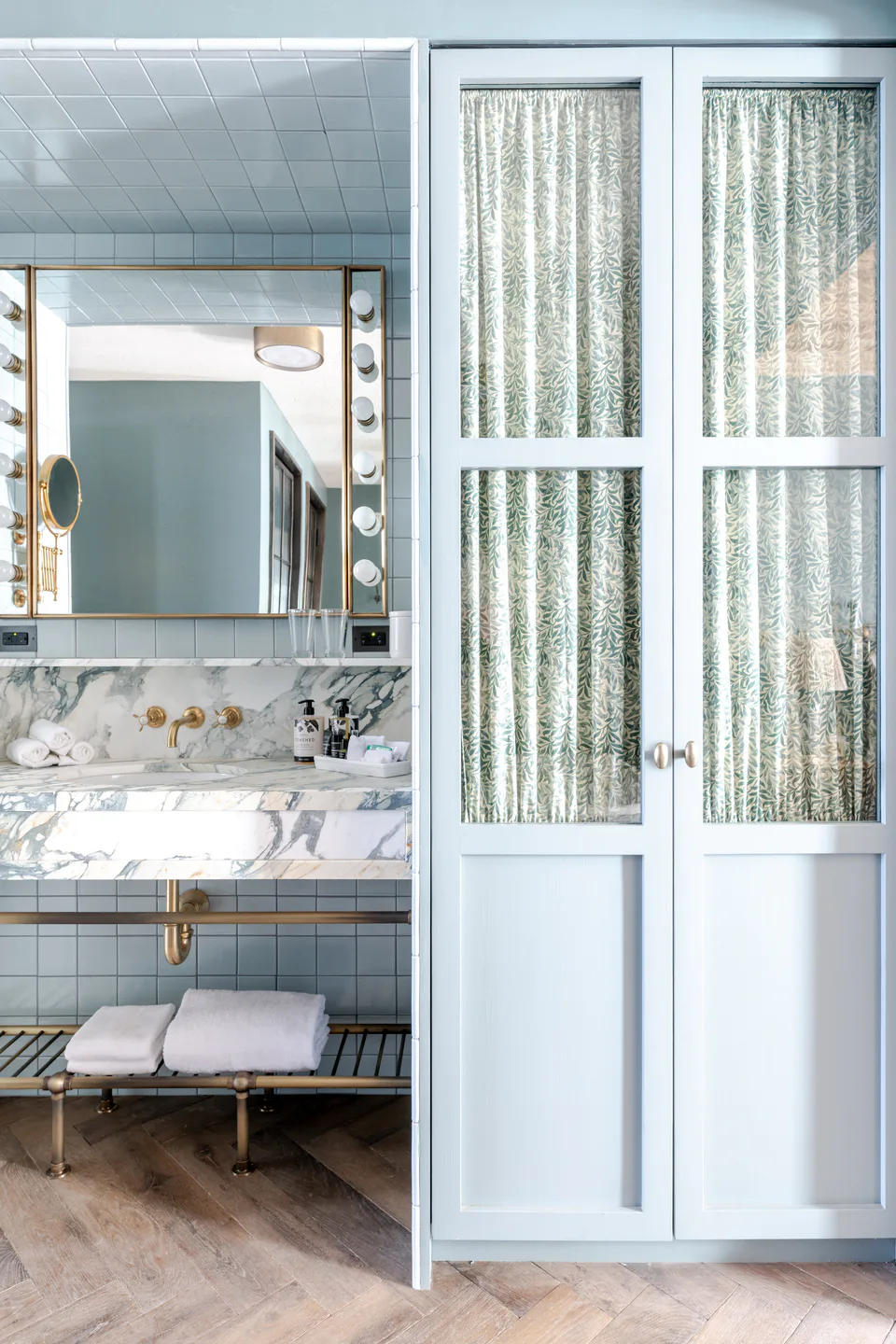 Medium bathroom with a marble vanity, a bulb-lit mirror and pale blue fabric-lined doors at Soho House Nashville, Nashville, Tennessee, interior design by Alexandra Peck Design